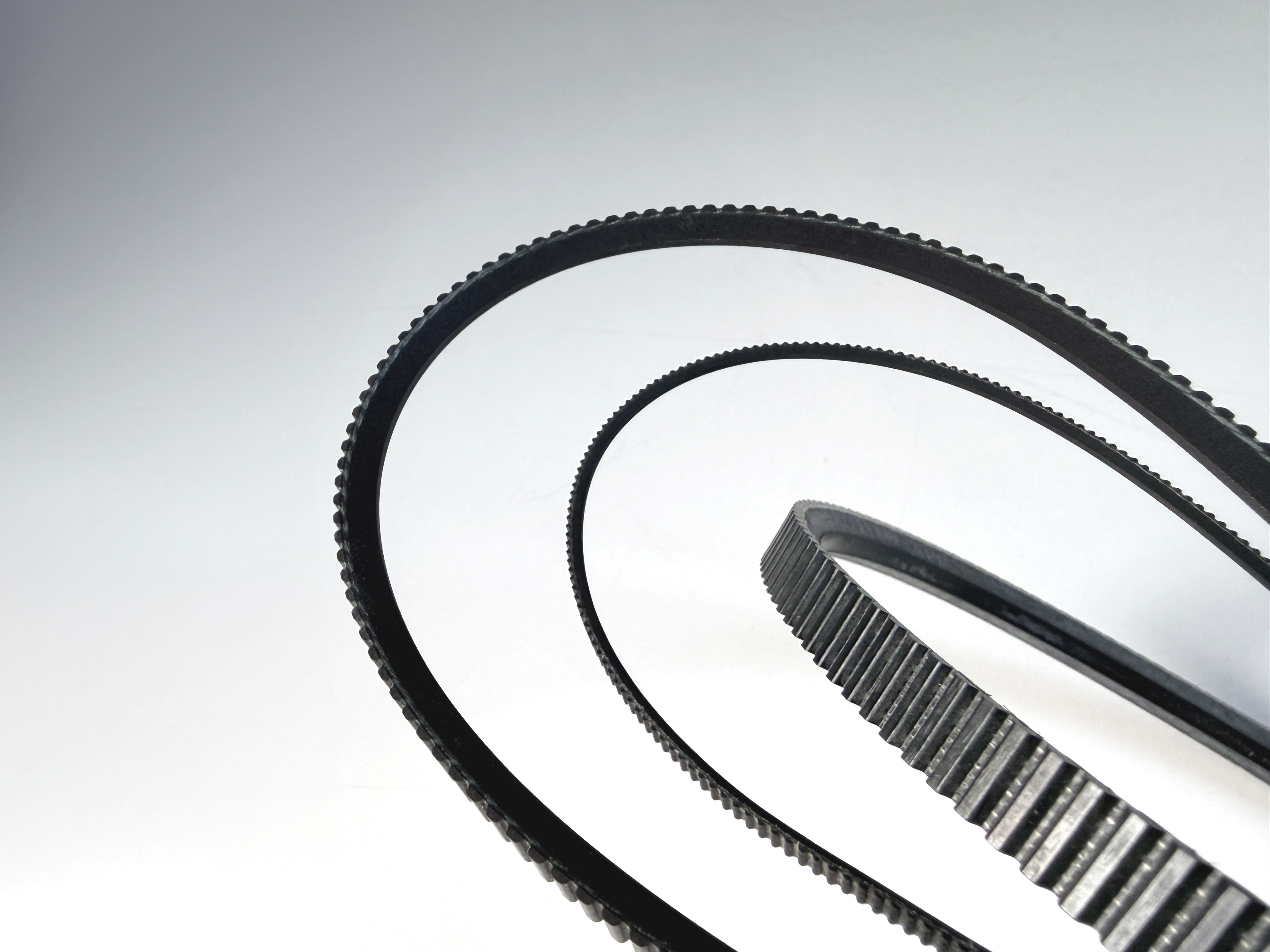 Mehrere Weitwinkelriemen mit unterschiedlichem Zahnprofil in geschwungener Form vor hellem Hintergrund